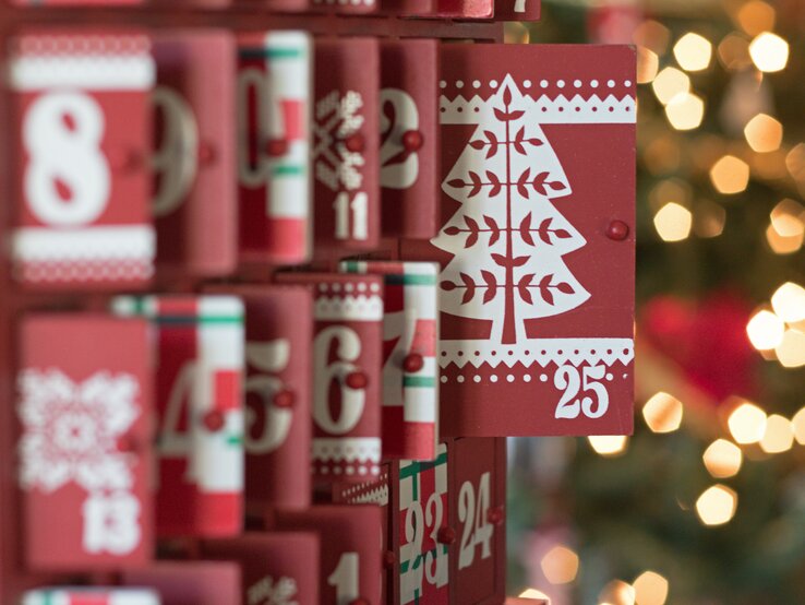 Roter Adventskalender mit weißen Zahlen und Weihnachtsbaum-Motiv auf Tür 25, weitere Türen teilweise geöffnet.