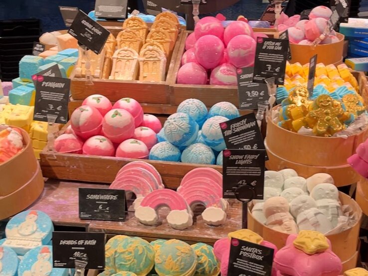 Farbige Badebomben und Seifen in verschiedenen Formen wie Regenbogen, Schneemänner und Lebkuchenmänner, dekorativ auf Holzkisten präsentiert.