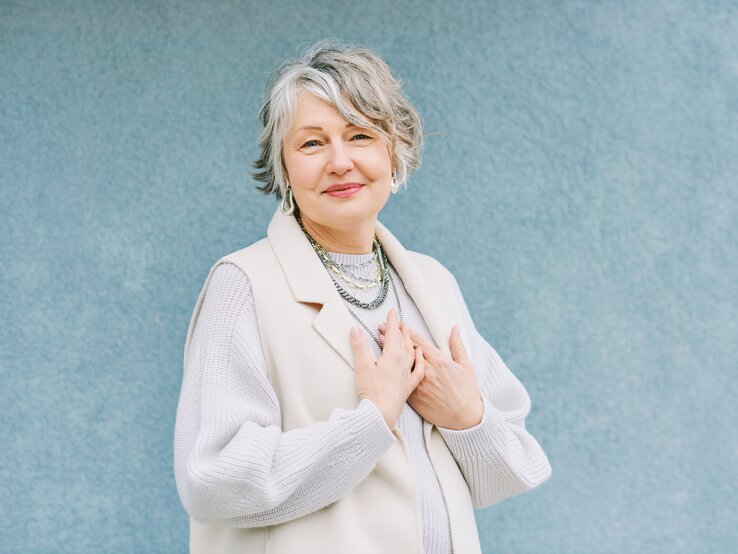 Eine lächelnde ältere Frau mit grauen Haaren, die eine elegante weiße Weste und einen gestrickten Pullover trägt, hält ihre Hände liebevoll auf der Brust. Sie steht vor einem einfarbigen blauen Hintergrund, der ein Gefühl von Ruhe und Gelassenheit vermittelt.