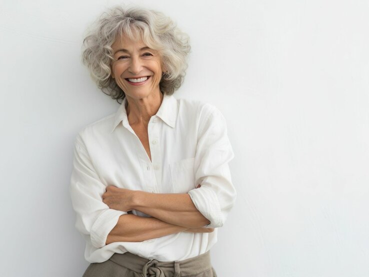 Eine fröhliche ältere Frau mit lockigem grauem Haar, die ein strahlendes Lächeln zeigt, steht vor einem weißen Hintergrund. Sie trägt ein klassisches weißes Hemd und eine beige Hose mit einem gebundenen Gürtel. Die Frau hat die Arme verschränkt und strahlt Selbstsicherheit und Zufriedenheit aus.
