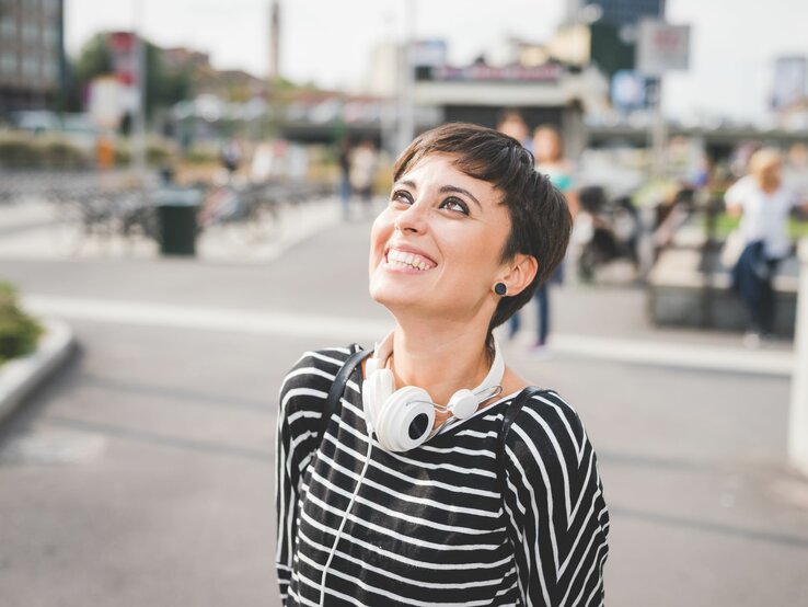 Lächelnde Frau mit einem kurzen Pixie Cut, die draußen steht. Sie trägt ein gestreiftes Oberteil und hat Kopfhörer um den Hals. Ihr Blick ist nach oben gerichtet
