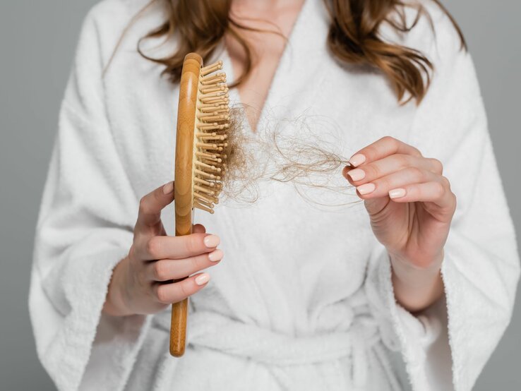 Frau im Bademantel, die besorgt eine Haarbürste mit ausgefallenen Haaren betrachtet. 