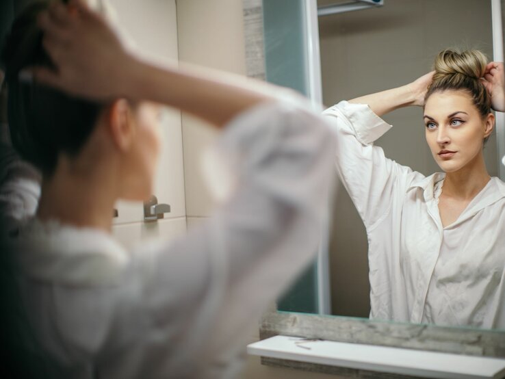 Frau, die sich in einem Spiegel betrachtet, während sie ihr Haar zu einem Dutt bindet. Sie trägt eine weiße Bluse