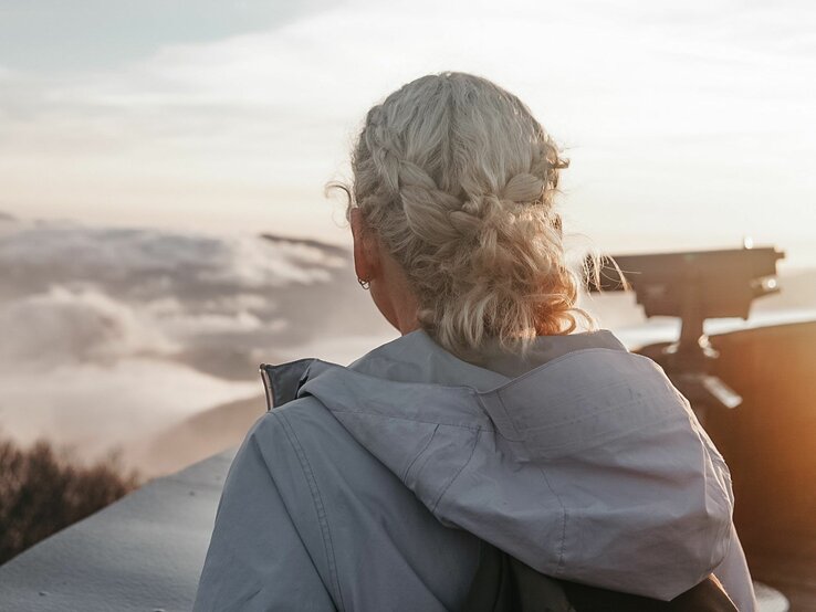 Junge Frau mit grauen, eingeflochtenen Haaren von hinten, die in die Ferne auf eine beeindruckende Berglandschaft blickt.