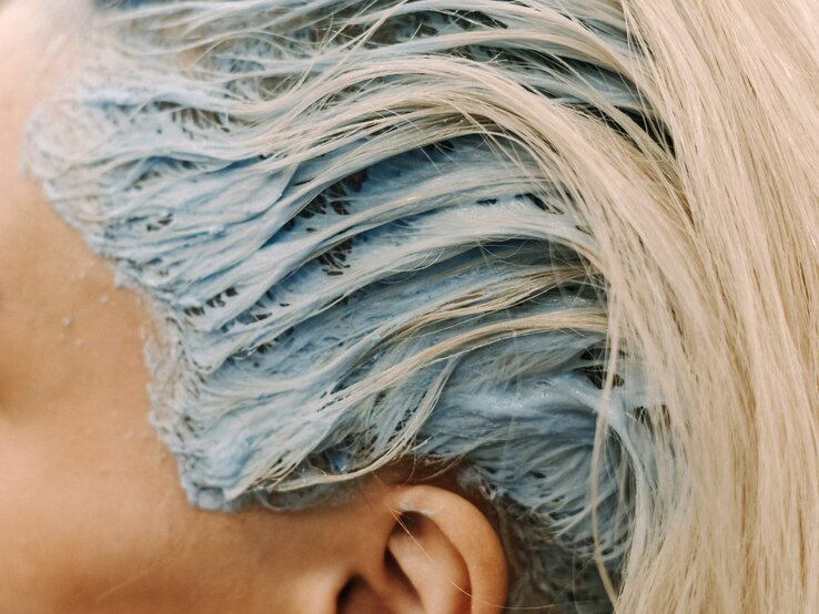 Detail einer Haarfärbung im Prozess. Die blonden Haare sind mit einer blauen Färbemasse bedeckt. Man kann die Struktur der Haare und die Cremigkeit der Farbe erkennen, die einen Kontrast zum natürlichen Blond der Haare bildet.