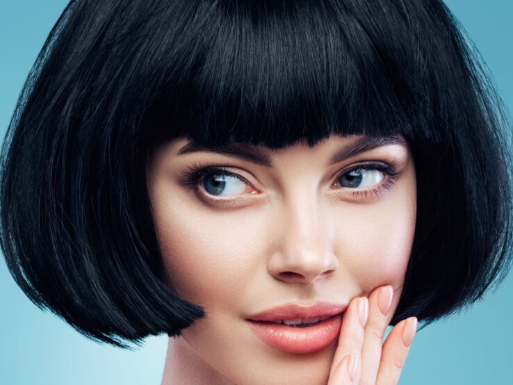 Frau mit einem schwarzen Bob-Haarschnitt und einem blauen Hintergrund. Sie hat blaue Augen und schaut leicht zur Seite, während sie ihre rechte Hand sanft an ihre Wange legt.