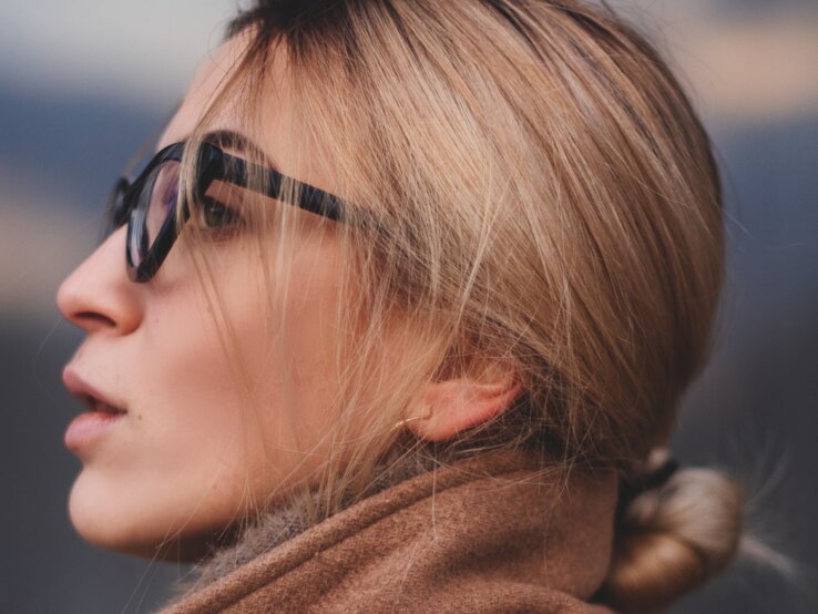 Junge Frau, die eine Sonnenbrille trägt und nachdenklich in die Ferne blickt. Sie hat blonde Haare, die zu einem lockeren Pferdeschwanz gebunden sind.