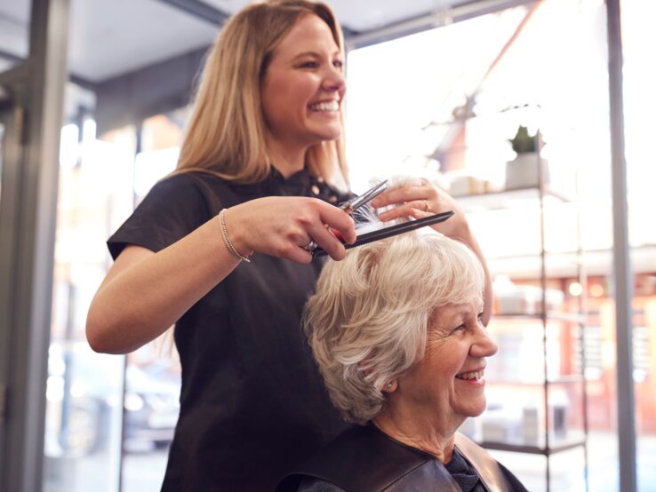 Eine junge Friseurin mit langen blonden Haaren schneidet lächelnd die Haare einer fröhlichen älteren Dame in einem hellen Salon.