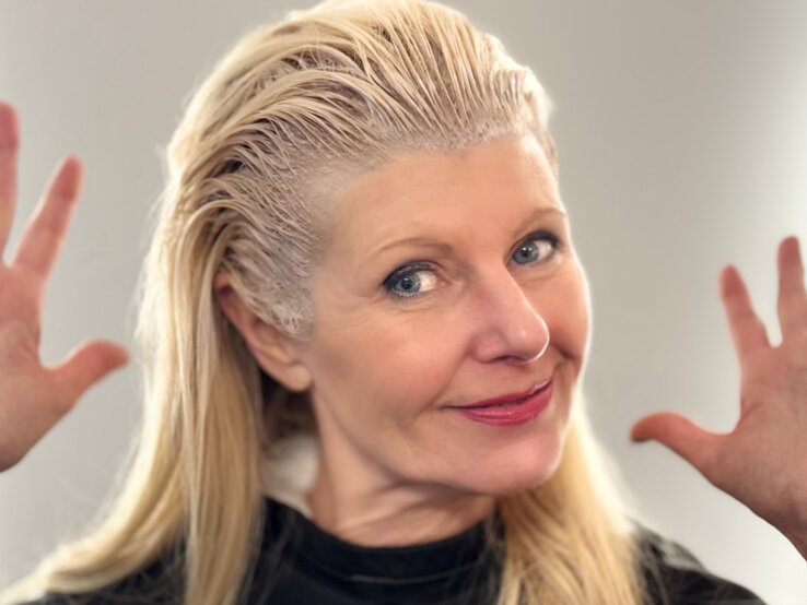 Renate Zott beim graue Haare färben, lächelt in die Kamera und hebt fröhlich die Hände.