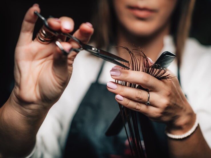 Nahaufnahme von Händen einer Friseurin mit Schere und Kamm beim präzisen Schneiden brauner Haare. Trendfrisuren 2025