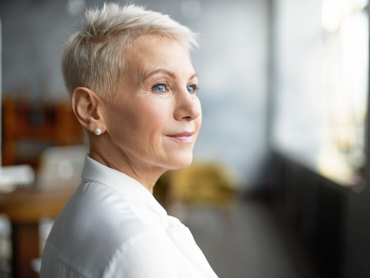 Reife Frau mit stylischer Pixie-Frisur - einem Kurzhaarschnitt für dünnes Haar - und Perlenohrringen trägt weiße Bluse und schaut verträumt ins Tageslicht.