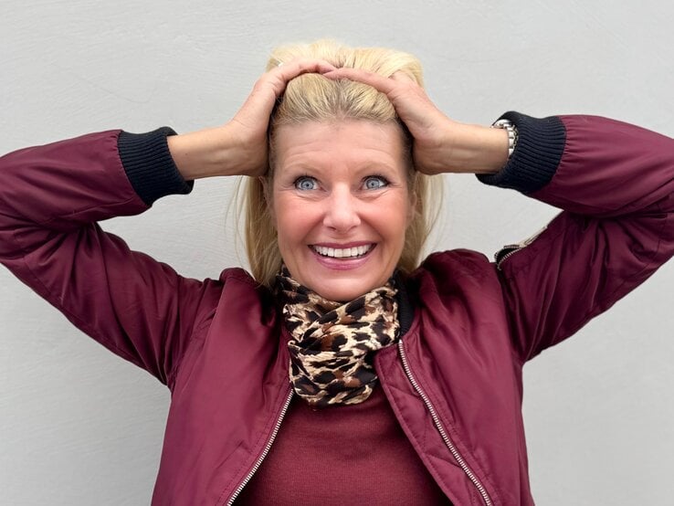 Renate Zott Bad Hair Day Sommer Fröhliche, blondhaarige Renate Zott, mittleren Alters hält sich mit beiden Händen die Haare, trägt bordeauxrote Jacke und Leoparden-Schal.