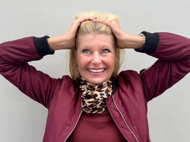 Renate Zott Haarpflege Fehler Sommer Fröhliche, blondhaarige Renate Zott, mittleren Alters hält sich mit beiden Händen die Haare, trägt bordeauxrote Jacke und Leoparden-Schal.