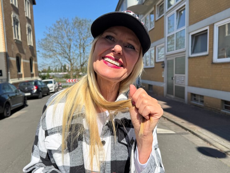 Renate Zott Bad Hair Day Sommer Cap | © Renate Zott Lachende Renate Zott mit Cap, pinkem Lippenstift und kariertem Mantel steht auf sonniger Straße zwischen Alt- und Neubauten. | © Renate Zott