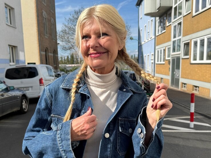 Renate Zott Bad Hair Day Sommer geflochtenes Haar | © Renate Zott Renate Zott mit geflochtenen Zöpfen, blauer Jeansjacke und hellen Lippen lächelt sanft bei Sonnenschein zwischen Wohnhäusern. | © Renate Zott