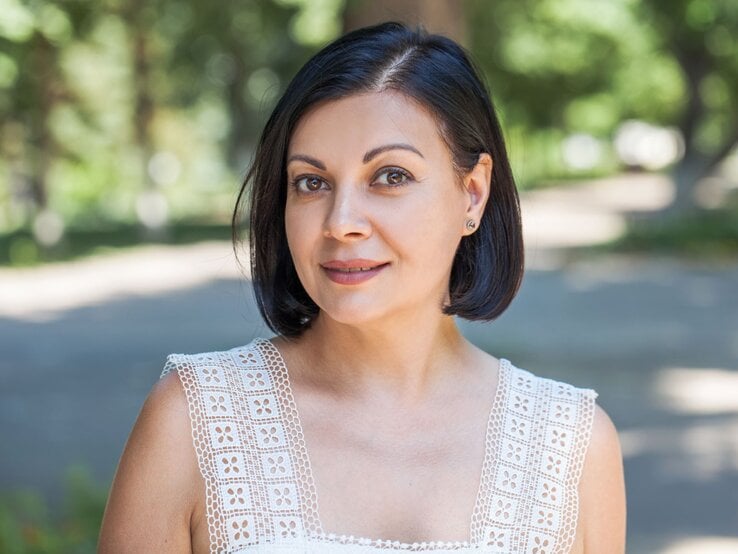 Brünette Frau mit kurzem, elegantem Grown-out-Bob und dezentem Make-up steht leicht lächelnd im Schatten eines grünen Parks.