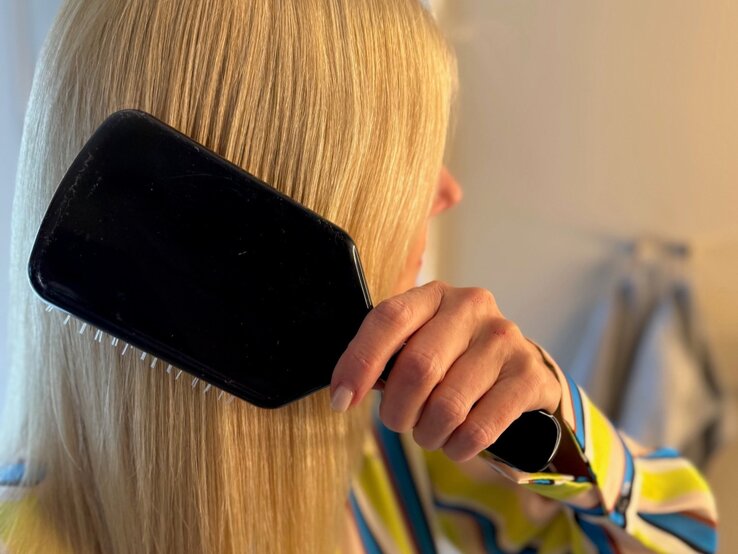 Renate Zott Haarpflege Fehler Bürste | © Renate Zott Renate Zott bürstet sich mit einer schwarzen Paddle-Brush die glatten Haare vor hellem Badezimmerhintergrund. | © Renate Zott