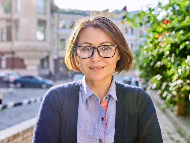 Frisuren für Frauen ab 50 mit Brille Freundlich blickende Frau mit braunem Bob und schwarzer Brille steht bei Sonnenschein in einer ruhigen Stadtstraße.