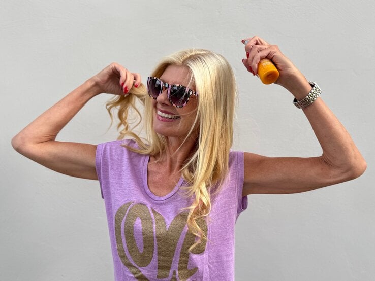 Renate Zott mit langen blonden Haaren sprüht sich mit Sonnenspray ein, trägt lila Top und bunte Sonnenbrille.