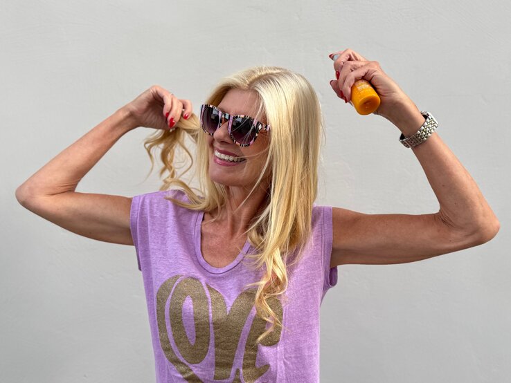 Renate Zott mit langen blonden Haaren sprüht sich mit Sonnenspray ein, trägt lila Top und bunte Sonnenbrille.