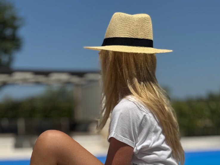 Renate Zott Haare Sonnenschutz Strohhut | © Renate Zott Renate Zott mit Strohhut und weißem T-Shirt sitzt am Pool und blickt in einen sonnigen Garten mit blauem Himmel. | © Renate Zott