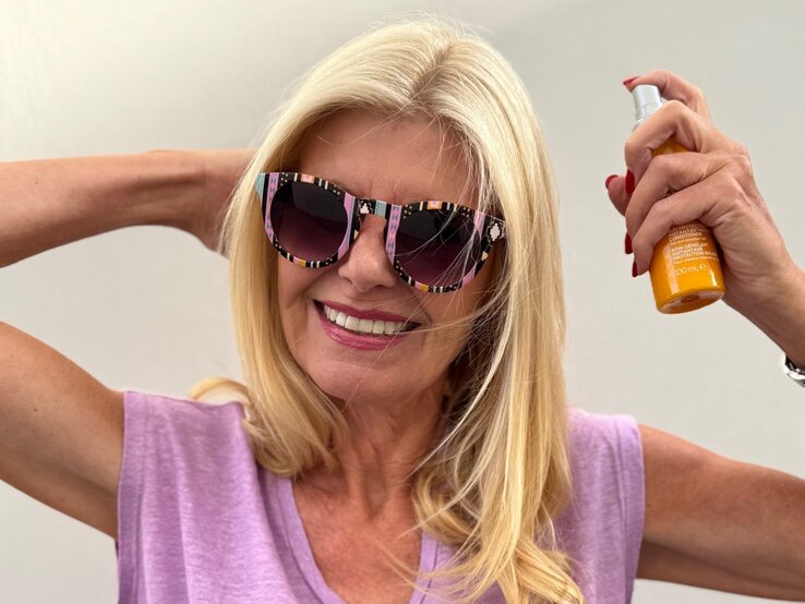 Renate Zott Sonnenschutz Haare Sommer Spray | © Renate Zott Renate Zott mit offenem blondem Haar posiert in pinker Sonnenbrille und lila Shirt, hält Sprayflasche hoch. | © Renate Zott