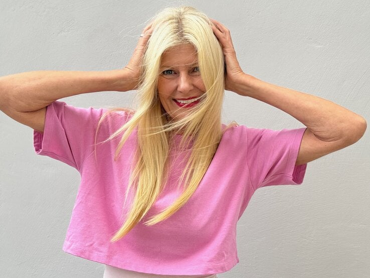 Renate Zott mit langen blonden Haaren und pinkem T-Shirt lächelt lebhaft, hält sich die Haare vor hellgrauer Wand.