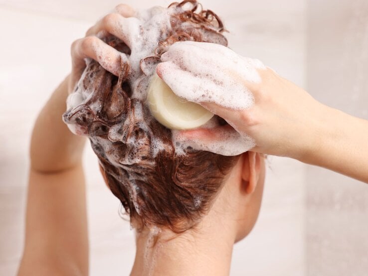 Eine Frau mit nassem, braunem Haar schäumt unter der Dusche mit fester Shampooseife kräftig den Kopf ein.