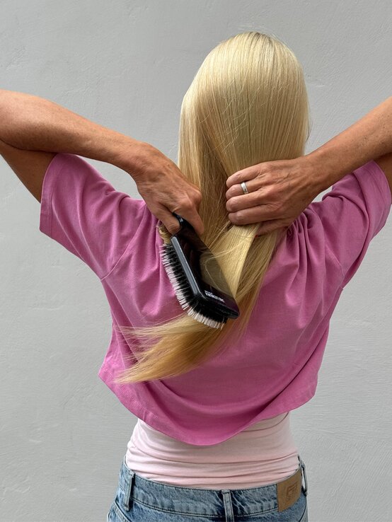Renate Zott in pinkem Shirt bürstet ihr glattes, langes Haar mit einer schwarzen Bürste vor grauer Wand. | © Renate Zott