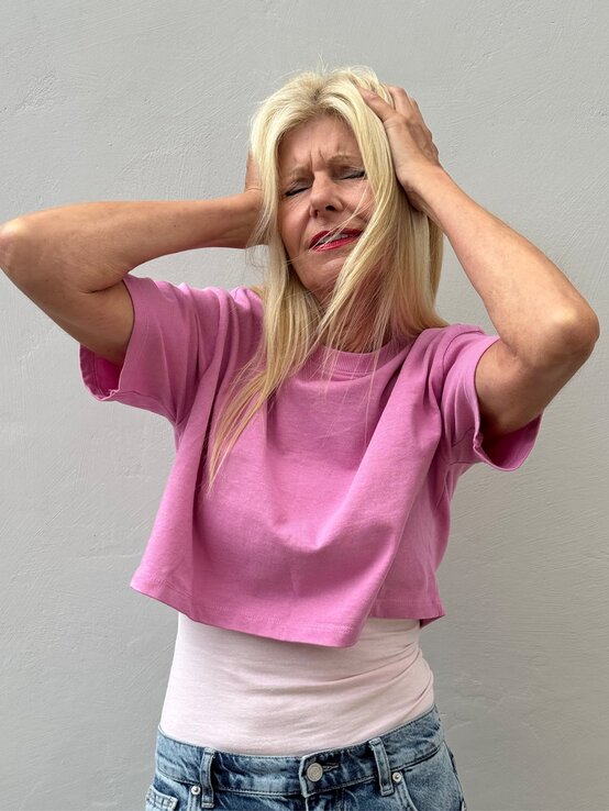 Renate Zott in pinkem Shirt hält sich mit schmerzverzerrtem Gesicht und geschlossenen Augen den Kopf. | © Renate Zott