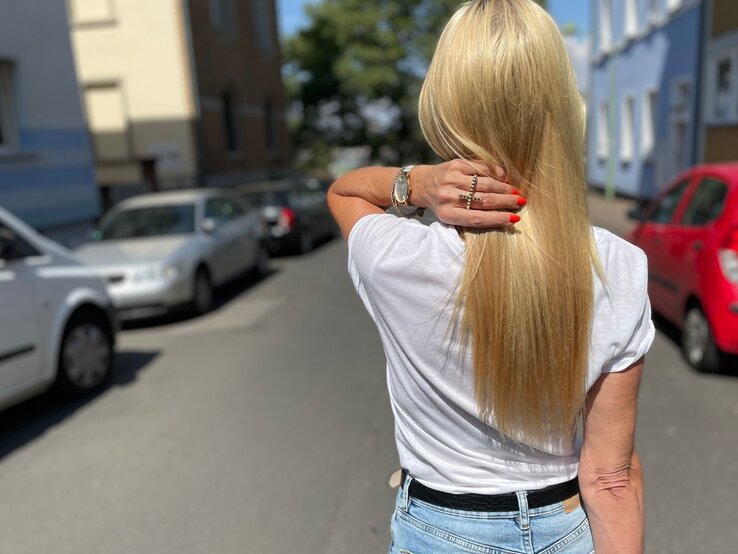 Eine Frau mit glattem, langen, blonden Haar, rotem Nagellack und Jeans streicht sich das Haar in einer ruhigen Wohnstraße zurück.