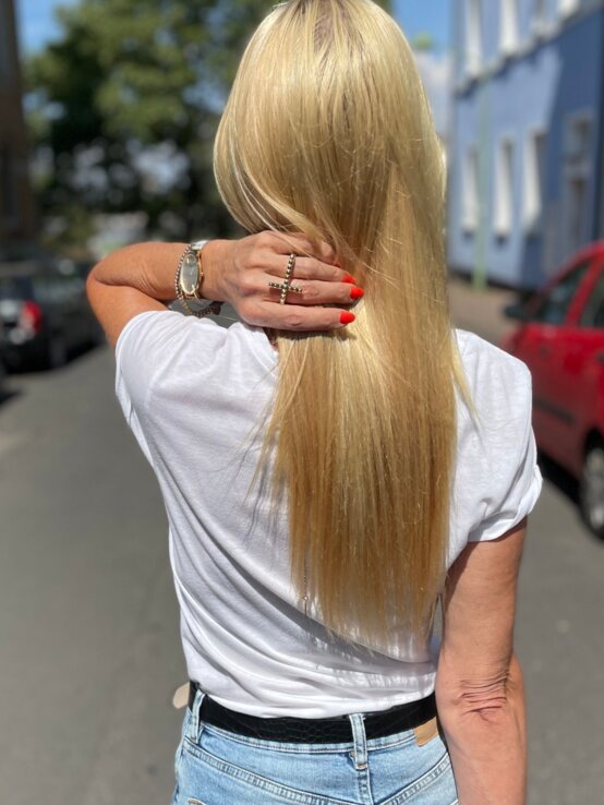 Eine Frau mit glattem, langen, blonden Haar, rotem Nagellack und Jeans streicht sich das Haar in einer ruhigen Wohnstraße zurück. | © Renate Zott
