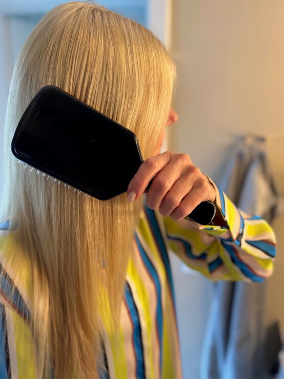 Detailaufnahme einer blonden Frau, die sich vor einem neutralen Hintergrund mit einer Bürste in der Hand die Haare kämmt. | © Renate Zott