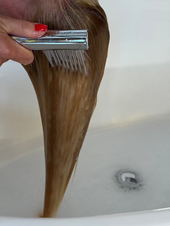 Eine Hand mit rotem Nagellack wäscht nasses, glattes Haar mit einem silbernen Duschkopf über einer mit Wasser gefüllten Badewanne. | © Renate Zott