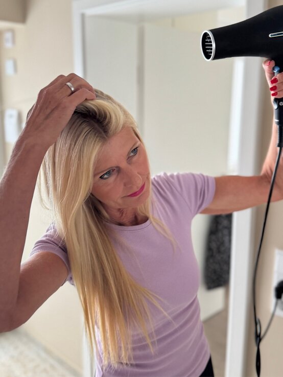 Blonde Frau im engen lila Oberteil verwendet einen schwarzen Föhn, während sie mit der Hand ihr Haar anhebt, in einem hellen Raum. | © Renate Zott