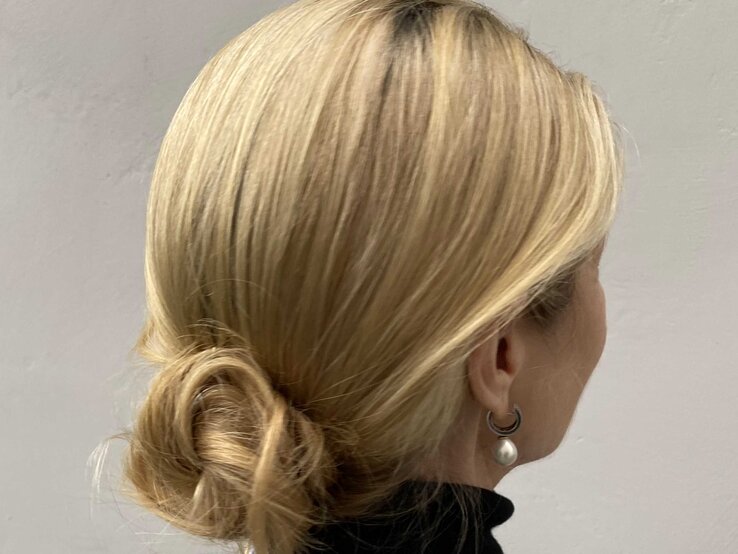 Eine Frau mit blondem Haar trägt eine elegante tief gebundene Frisur und Perlenohrringe vor einer schlichten, hellgrauen Wand. | © Renate Zott 