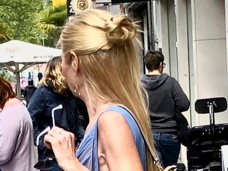 Frau mit langen blonden Haaren, teilweise hochgesteckt, trägt ein hellblaues Kleid und Sonnenbrillen in der Hand, in einer belebten Straße. | © Renate Zott 