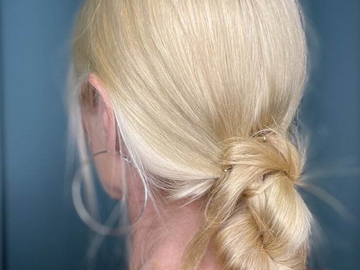 Kunstvoll geflochtene blonde Frisur mit dezenten goldenen Haaraccessoires, kombiniert mit schlichten silbernen Ohrringen und blauem Hintergrund. | © Renate Zott 