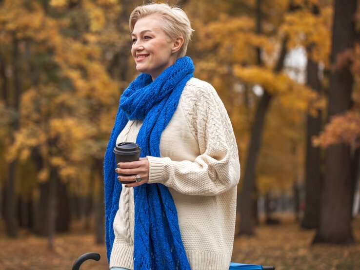 Person mit kurzem blondem Haar trägt cremefarbenen Strickpullover, blauen Schal und hält einen Kaffeebecher im herbstlich bunten Park.