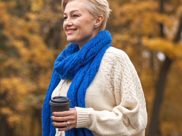 Person mit kurzem blondem Haar trägt cremefarbenen Strickpullover, blauen Schal und hält einen Kaffeebecher im herbstlich bunten Park.