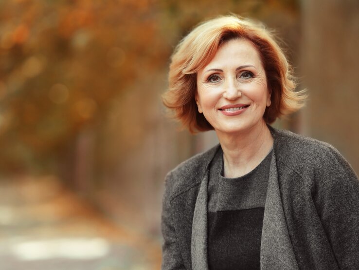Eine Frau mit schulterlangem rotblondem Haar trägt einen grauen Mantel und steht vor einem herbstlich verschwommenen Hintergrund.