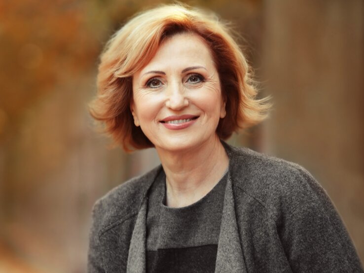 Eine Frau mit schulterlangem rotblondem Haar trägt einen grauen Mantel und steht vor einem herbstlich verschwommenen Hintergrund.
