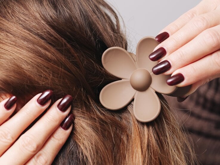 Zwei Hände mit bordeauxroten Fingernägeln klemmen eine braune Haarspange in Blumen-Form in braunes Haar.
