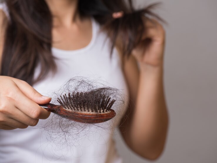 Dunkelhaarige Frau im weißen Top hält besorgt eine Haarbürste voller Haare, während sie eine Haarsträhne prüfend betrachtet.