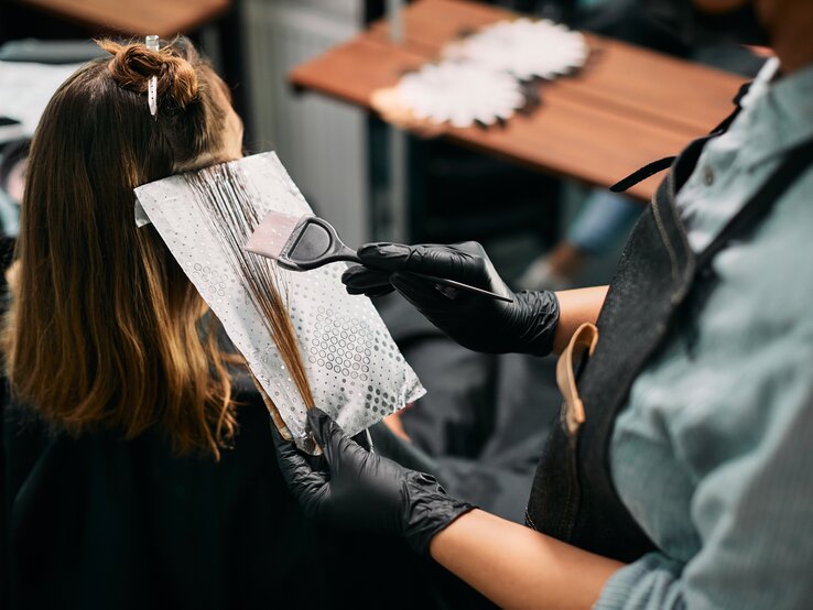 Friseur mit schwarzem Handschuh trägt mit Pinsel Haarfarbe auf braune Strähnen einer Frau auf, die in einem Salon sitzt.