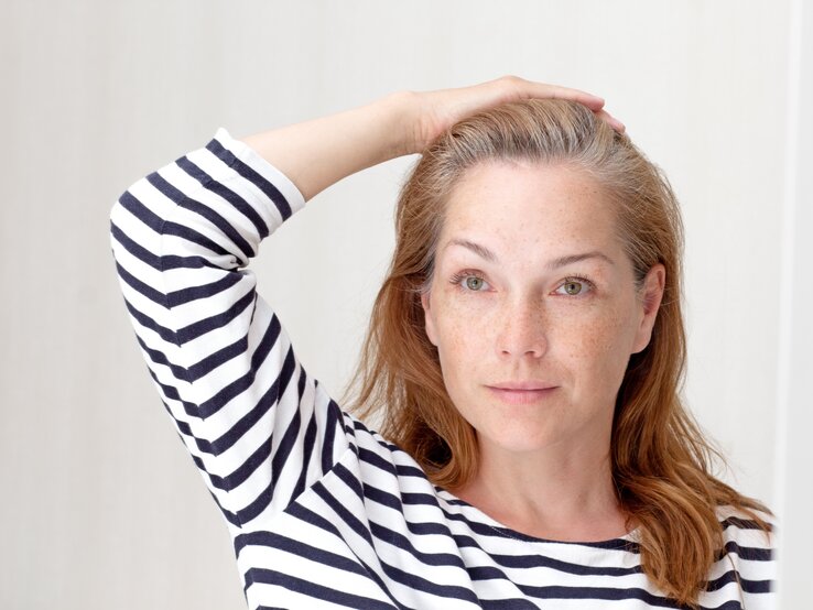 Eine Person mit halblangem rotbraunem Haar und grauem Ansatz trägt ein gestreiftes Shirt und hält eine Hand auf den Kopf vor hellem Hintergrund.
