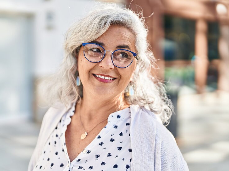 Graues Haar in leichten Wellen, weiß-blau gemustertes Hemd mit goldener Halskette und hellem Cardigan, Hintergrund unscharf.