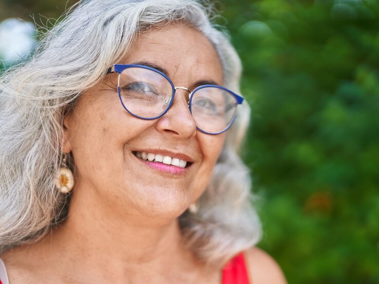 Frau mit grauem Haar, baumelnden Muschel-Ohrringen, einer blauen Brille und rotem Oberteil, umgeben von grünem, unscharfem Naturhintergrund.
