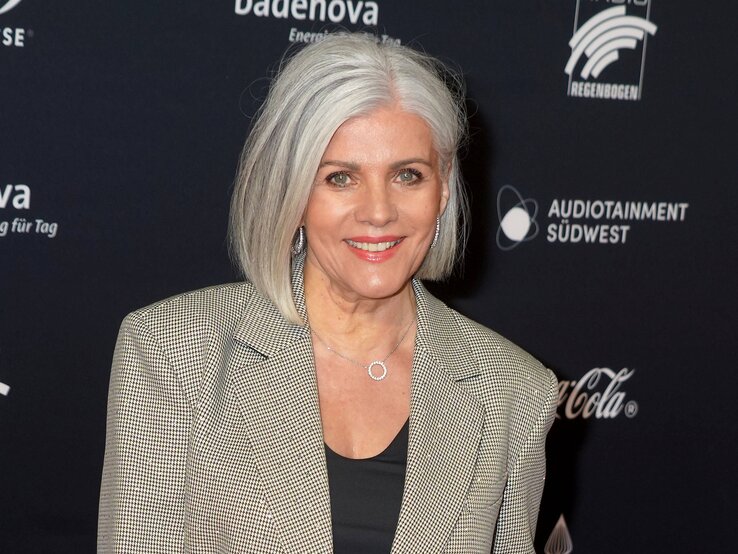 Birgit Schrowange mit schulterlangem grauem Haar trägt karierten Blazer und schwarzen Pullover, steht vor schwarzem Hintergrund mit weißen Logos.