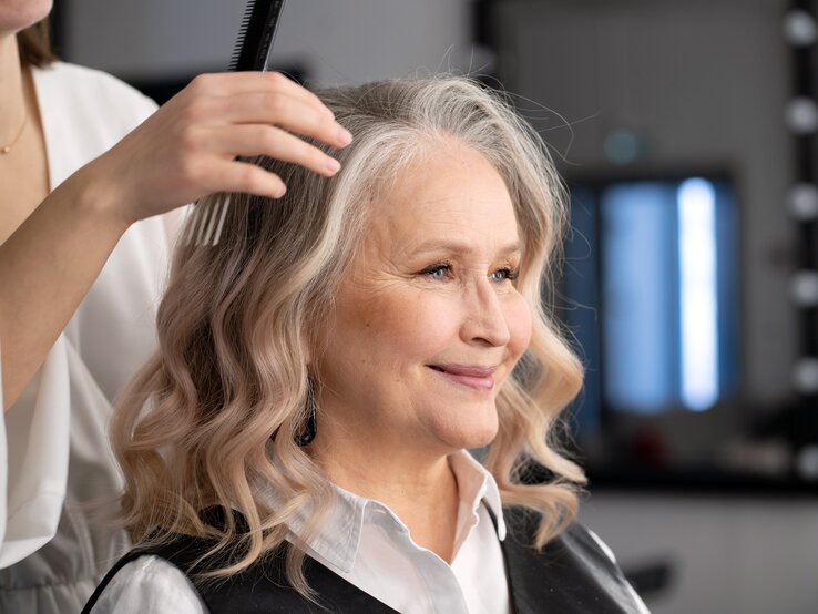 Eine Hand im weißen Ärmel kämmt weiche, blonde Locken; im Hintergrund ein dunkel eingerichteter Friseursalon.